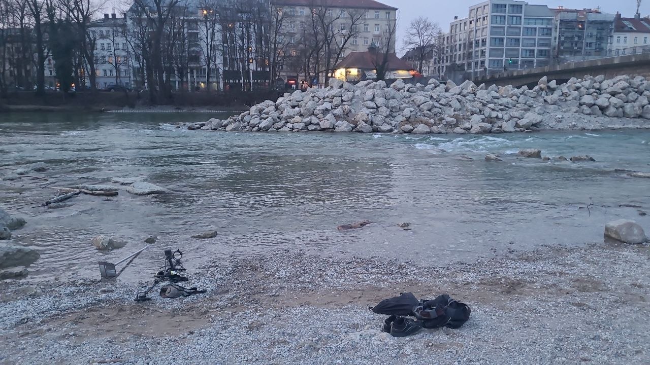 Zwei Männer stehen am steinigen Ufer der Isar in München, einer hält einen Ring in die Höhe, im Hintergrund eine Kirche bei Abendlicht.